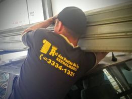 Pessoa ajustando porta de aço em garagem, com uniforme de serralheria.