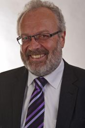 Mann mit Brille und grauem Bart im Anzug, lächelnd, vor neutralem Hintergrund.