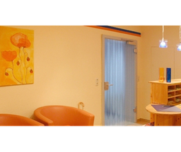 Zwei braune Stühle in einem Wartezimmer, daneben ein gelbes Bild mit roten Blumen an der Wand.