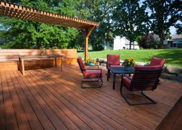 Wooden deck with pergola, red cushioned chairs, tables, and bench, surrounded by greenery.