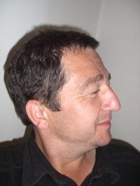 Ein Mann im Profil mit dunklem Haar, der nach rechts schaut, vor einer weißen Wand.