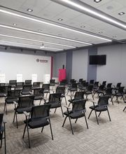 Sala de conferências vazia, cadeiras pretas organizadas em fileiras e telões nas paredes cinzas.