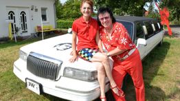Frau sitzt auf Limousine, Mann in buntem Outfit daneben. Haus und Bäume im Hintergrund.