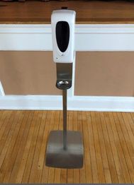 Touchless hand sanitizer dispenser on a metal stand in a room with wooden floors.