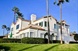 White house with red roof tiles, surrounded by palm trees and green grass under a clear blue sky.