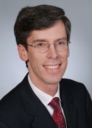 Porträt eines lächelnden Mannes mit Brille und Anzug mit roter Krawatte.