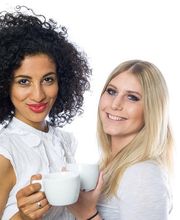 Zwei Frauen mit Kaffeetassen lächeln in die Kamera. Hintergrund ist weiß.
