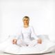 Frau meditiert im Schneidersitz auf Kissen, trägt weiße Kleidung, vor weißem Hintergrund.