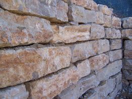 Muro de piedra seca hecho de bloques irregulares, con textura rugosa y color marrón claro.