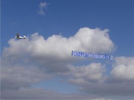 Ein Flugzeug zieht ein Banner mit der Aufschrift "sky-luftwerbung.de" am Himmel vor Wolken.