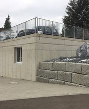 Haus mit Betongarage und steiniger Landschaft, oben sind geparkte Autos sichtbar.