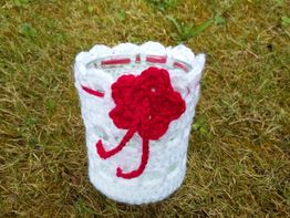 Glas mit weißer Häkelhülle und roter Blume, stehend auf einer Wiese.