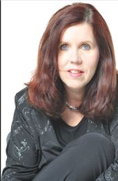 Frau mit schulterlangem braunem Haar und blauem Blick, trägt schwarze Kleidung, sitzt vor weißem Hintergrund.