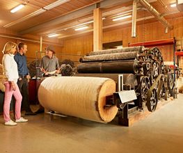 Drei Personen betrachten in einer Werkstatt eine große alte Textilmaschine mit Wollbahnen.