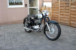 Klassisches Motorrad im Hof, geparkt auf gepflastertem Boden vor einem Holzzaun und einem Haus.