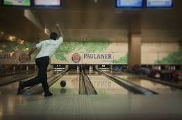 Persona lanzando una bola de bolos en una bolera, fondo con cartel "Paulaner".
