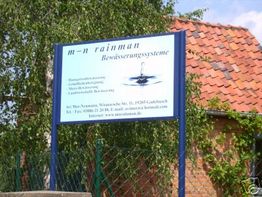 Schild mit der Aufschrift "m-n rainman Bewässerungssysteme" vor Ziegelgebäude und Baum.