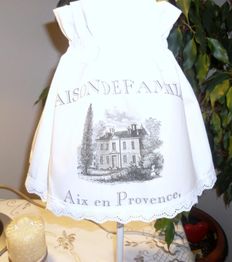 Tischlampe mit weißem Überzug und Aufschrift "Maison de Famille, Aix en Provence" neben Pflanze.
