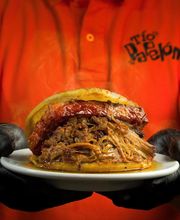 Persona con guantes negros sostiene una hamburguesa grande en un plato.