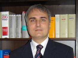 Ein Mann im Anzug steht vor einem Bücherregal mit verschiedenen Gesetzbüchern.