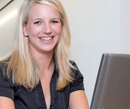 Eine lächelnde Frau mit blondem Haar sitzt am Laptop in einem Büro.