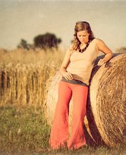 Frau lehnt an Heuballen auf Feld, trägt rote Hose, blondes Haar, friedliche Landschaft.