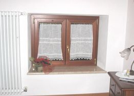 Finestra con tende in pizzo bianco, termosifone, pianta e lampada su mobile vicino.