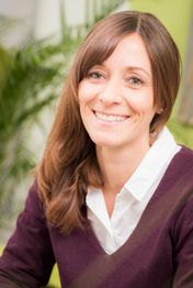 Eine lächelnde Frau mit braunem Haar vor einem verschwommenen, grünen Hintergrund.