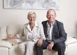 Ein lächelndes Paar sitzt auf einem hellen Sofa, sie tragen elegante, helle Anzüge.