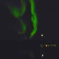 Grüne Nordlichter leuchten am Nachthimmel über einem See mit Lichtern am Ufer.