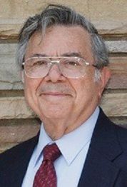 Elderly person with glasses in a suit, standing against a stone wall, smiling slightly.