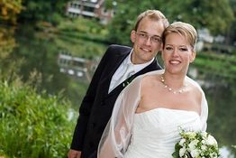 Ein Hochzeitspaar posiert lächelnd am Ufer eines Flusses vor einer Kulisse aus Bäumen und Häusern.