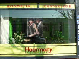 Haarstudio-Fenster mit Werbung für Kosmetik und Fußpflege. Im Inneren sind Pflanzen sichtbar.