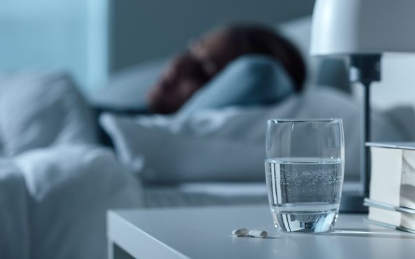 Glas Wasser und Melatonin-Tabletten auf einem Nachttisch vor einer schlafenden Person.
