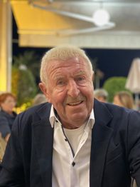 Älterer Mann in einem Anzug sitzt lächelnd im Freien bei Nacht.