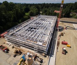 Bau einer großen Halle mit Stahlkonstruktion, umgeben von Baugeräten und Bäumen im Hintergrund.