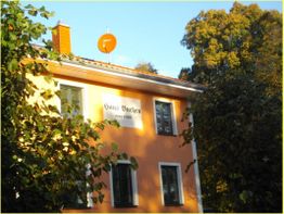Orange Haus "Hans Buchen" mit Sat-Schüssel, umgeben von Bäumen bei Sonnenschein.