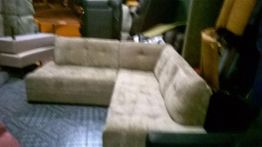 Sofá de canto bege em ambiente interno com piso escuro e vários objetos ao redor.