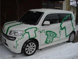 Weißes Auto mit grünen T-Shirt-Motiven und Druckschrift auf schneebedecktem Boden.