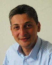 Eine lächelnde Person mit kurzem, grauem Haar in einem gestreiften Hemd vor weißem Hintergrund.