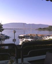 Blick auf einen ruhigen Hafen mit Booten und Bergen im Hintergrund, aufgenommen von einer Terrasse.