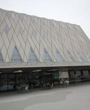 Modernes Gebäude mit schräger Fassade und dreieckigen Fenstern, Menschen sitzen draußen.