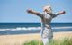 Frau mit grauen Haaren steht mit ausgebreiteten Armen am Strand, schaut aufs Meer.