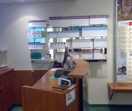 Opticien interieur met balie, computer, brillen bij display, en dozen met lenzen op planken.