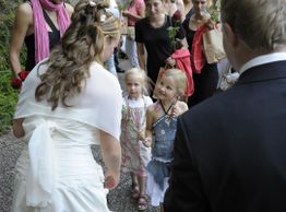 Braut und Gäste stehen im Freien. Zwei kleine Mädchen halten Rosen. Feierstimmung herrscht.