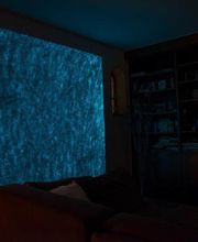 Dunkles Wohnzimmer mit beleuchteter blauer LED-Wand und braunem Sofa im Vordergrund.