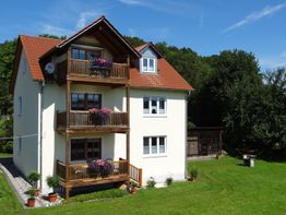 Zweistöckiges Haus mit rotem Dach, Balkonen und Garten, umgeben von Bäumen und blauem Himmel.