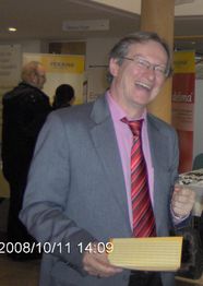 Ein lächelnder Mann im Anzug hält ein gelbes Buch. Im Hintergrund sind Plakate und Menschen.