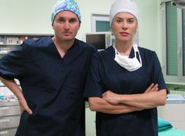 Due medici in sala operatoria, indossano uniformi blu e cuffie chirurgiche.