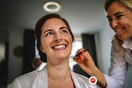 Vrouw lacht terwijl ze visagie krijgt in een lichte kamer. Visagiste houdt een penseel vast.